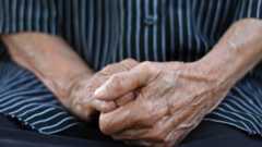 Close-up of the hands of an elderly person that are crossed together over their lap. They are wearing a shirt with black and light blue stripes. 