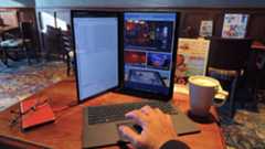 A dual-screen laptop is set up in a vertical orientation with the detachable keyboard in front, and a hand is shown resting on the trackpad.