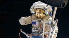 an astronaut in space, his golden visor reflecting a bent image of a space station module out of frame, gives a thumbs up. In the foreground immediately in front of him, a contraption of metal rods, similar to a folded camera tripod, extends the height of the image.