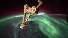 a folded robot arm in front of the earth and black space. the earth is in nighttime and green, swirling auroras are shining above the surface