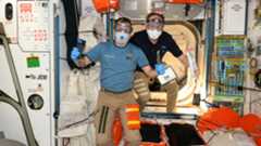 two men in short sleeve shirts and kahki slacks wear protective glasses, masks and gloves to protect against floating debris before entering a cargo ship berthed with a space station.