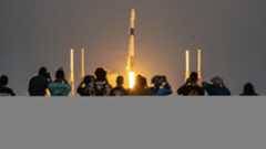 a row of people in the distance take photos of a rocket far away but large in frame.