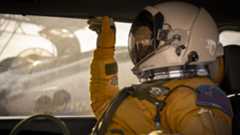 A person wears a tan astronaut suit and helmet and sits in the passenger seat of a car while a plane sits behind them on a tarmac.