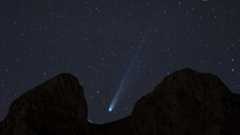 A white streak of a comet&#039;s tail can be seen in the center of the image as it streaks diagonally from top right to lower left between the dark silhouette of rocks blocking part of the starry night sky