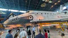 The space shuttle Discovery, a white and black shuttle is on display at the Smithsonian Museum where people walk around it and view it.