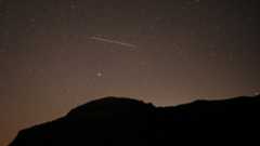 A lone meteor is spotted streaking through a dark starry sky above a silhouetted hill.