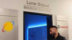 A man wearing a dark polo button up shirt heads into a blue-lit door with the words "Lunar Outpost: Mission Control" above it.