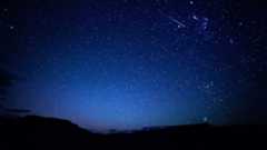 A star-studded sky glitters above an undulating, dark, silhouetted horizon. The sky is a lighter shade of blue towards the horizon, where the outlines of clouds can be seen hugging the skyline. A lone meteor is visible streaking Earthwards towards the upper left of the image.