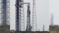 A white and black rocket is shown at a launch pad next to its launch tower and supports during the day, with a second large orange rocket with a white top and boosters pictured in the background.