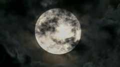 A full moon is shown against a black sky surrounded by clouds, with some encroaching on its surface.