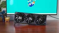 Palit GeForce RTX 5060 Ti Inifnity graphics card standing horizontally on woodgrain desk with monitor in backdrop with green wallpaper next to plant