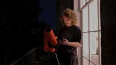 A man with long hair stood next to a bright window and red smart telescope at night whilst looking at a smartphone.