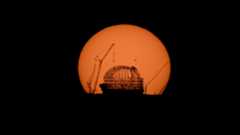 A large orange sun illuminates the scaffolding of a dome where a telescope is being built, along with cranes next to it.