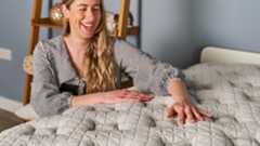 The image shows an woman smiling as she places her hands on the surface of a white mattress while she crouches next to it