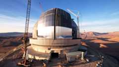 An enormous telescope dome is pictured in a desert during the day surrounded by cranes. Two huge silver sliding doors are visible, closed in the middle of the dome.