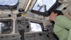 A woman wearing a headset and a green long sleeve shirt looks out of the windows on the International Space Station aimed at Earth