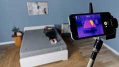 This image shows a behind the scenes of our temperature regulation testing. In the foreground is a phone attached to a rig, displaying a thermal image of our tester lying on a mattress. In the background, our sleep writer Ruth Jones lies on the Siena Signature Mattress on a bed frame in our Sleep Lab