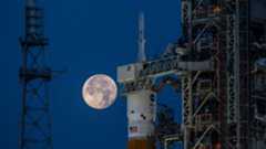 the full moon hangs in frame with the orion, inside its payload shell atop the SLS rocket.