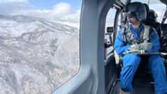 A man in a blue jumpsuit sits next to a plane window wearing a helmet and headset as he looks down over a series of snowy mountains