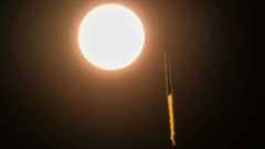 a rocket is seen in slihouette launching to space as it appears to pass by the sun 