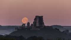 A castle ruin is in the foreground of the image with a subtle pink hued sky and a bright full moon.