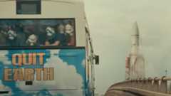 a large rocket looms in the distance behind a bus with the words "Quit Earth" on it