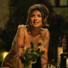 A woman (Emily Bader as Poppy) stands while resting both hands on an outdoor table decorated with wildflowers and bottles, in the Netflix movie 'People We Meet on Vacation.'