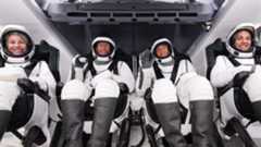 four astronauts in black and white spacesuits sit inside a space capsule