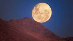 A glowing yellow moon is seen in a deep blue night sky with a red ridge of rock in front