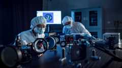 Two people wearing blue clean suits and masks bend over a laser table full of mirrors, lenses, and lasers
