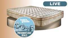 The Saatva Classic mattress shown on a gold plinth, with a blue Tom's Guide Presidents' Day deal badge overlaid on the bottom of the mattress. A LIVE badge is located in the top-right foreground. 