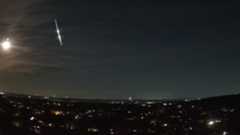  A bright fireball meteor is shown streaking earthward through the night sky over the lights of a town. The bright reflected light of the moon is visible to its left.