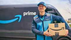 An Amazon Prime delivery driver in branded uniform holding packages while standing in front of a Prime-branded delivery van.