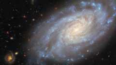spiral galaxy occupies most of the image. It is a slightly tilted disc of stars, yellow-white in the centre and blue in the outskirts, showing light from different stars in the galaxy. Its spiral arms curl outwards from the centre, speckled with blue star clusters. Dark reddish threads of dust swirl around the galaxy’s centre. The backdrop is two medium-sized and many small, distant galaxies on a black background.
