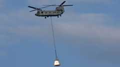 A helicopter lifts a large white crew module hoisted by a long rope in the air