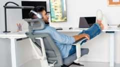 A man wearing headphones is relaxing in a light gray and white ergonomic office chair with his feet propped up on a white desk.