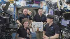 Four astronauts wearing black shirts float around a bag of prepackaged food in the International Space Station