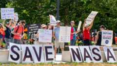 People hold signs outside on a sunny day in protest of NASA budget cuts, July 20, 2025.