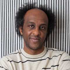 A man with short curly hair and a receding hairline is wearing a light sweater with black stripes. He is standing in front of a background with vertical black-and-white stripes, smiling softly with his arms crossed.