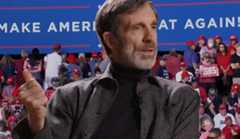 A white man in a check jacket speaking in front of a backdrop of a MAGA rally