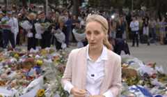A slender blonde woman stands in front of a floral memorial with mourners in the background.