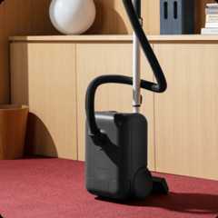 The sleek Airsign vacuum cleaner sits in a well-lit living room on red carpet, leaning against wood cabinets.