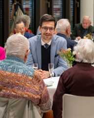 Dieser Alternativtext wurde KI-generiert: Mehrere Personen sitzen an einem festlich gedeckten Tisch und unterhalten sich. Blumen und Gläser mit Flüssigkeit sind auf dem Tisch zu sehen.