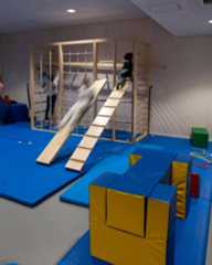 Dieser Alternativtext wurde KI-generiert: Ein Innenraum mit einer Spielstruktur aus Holz, bestehend aus einer Kletterwand und einer Rutsche. Der Raum ist mit blauen und gelben Matten ausgestattet.