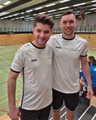 Dieser Alternativtext wurde KI-generiert: Zwei Personen in hellen Sporttrikots mit dunklen Akzenten stehen nebeneinander und schauen in die Kamera. Im Hintergrund ist eine Sporthalle mit einer bunten Wandgestaltung zu sehen.