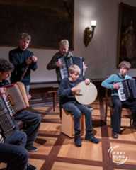 Dieser Alternativtext wurde KI-generiert: Eine Gruppe von Personen spielt verschiedene Musikinstrumente in einem Innenraum. Die Aufnahme ist farbig und zeigt eine Szene mit traditionellen Instrumenten.