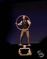 Dieser Alternativtext wurde KI-generiert: Eine Person steht auf einer schmalen Plattform und hält einen großen Kreis in der Luft. Der Hintergrund ist dunkel und das Bild wirkt wie eine Bühnenaufnahme.