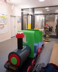 Dieser Alternativtext wurde KI-generiert: Ein Innenraum mit Holzwänden und einem großen Fenster. Im Vordergrund steht ein Spielzeugzug in den Farben Rot, Grün und Schwarz.