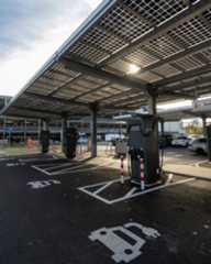 Dieser Alternativtext wurde KI-generiert: Ein überdachter Parkplatz mit Solaranlagen auf dem Dach und mehreren Ladestationen für Elektrofahrzeuge. Im Hintergrund sind weitere Fahrzeuge und eine Straße zu sehen.