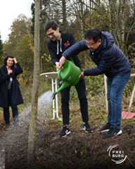 Dieser Alternativtext wurde KI-generiert: Mehrere Personen gießen einen jungen Baum im Freien. Im Hintergrund ist ein bewaldeter Hügel zu sehen.